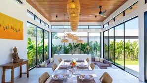 A modern dining area with a long table set for six, surrounded by large windows offering a view of lush greenery. - Villa Linaria by Pertama Stay (Ungasan)