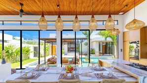 A dining area with a large table set for six, overlooking a pool and outdoor seating. - Villa Linaria by Pertama Stay (Ungasan)