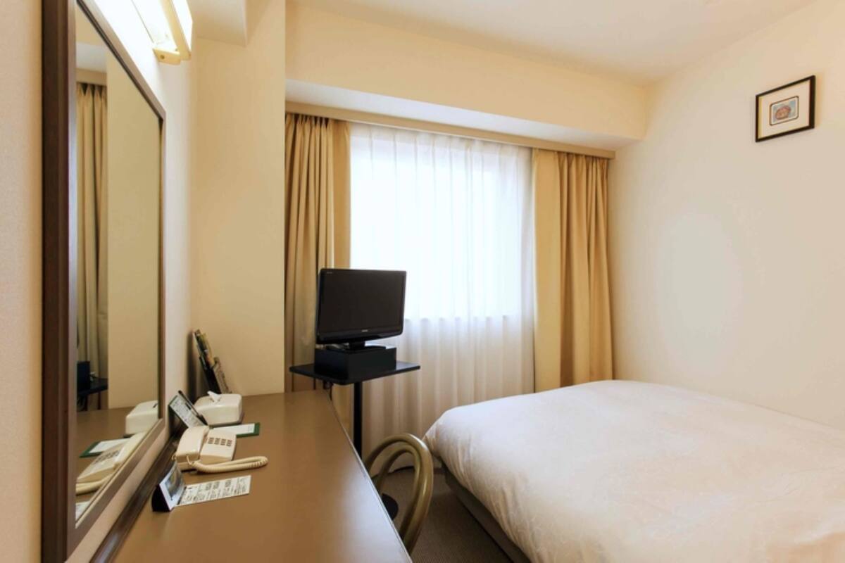 A hotel room with a bed, a desk with a telephone, a television, and a window with curtains.