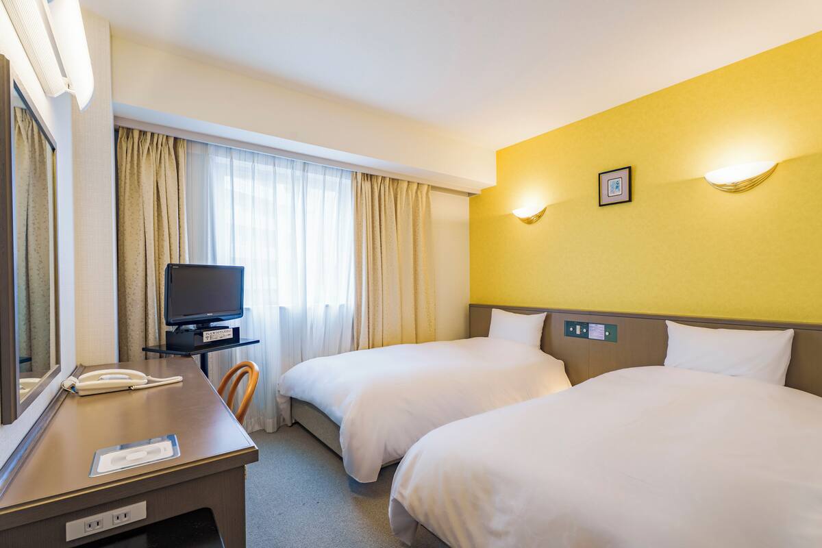 A hotel room with two beds, a desk with a telephone, a television, and a large window with curtains.