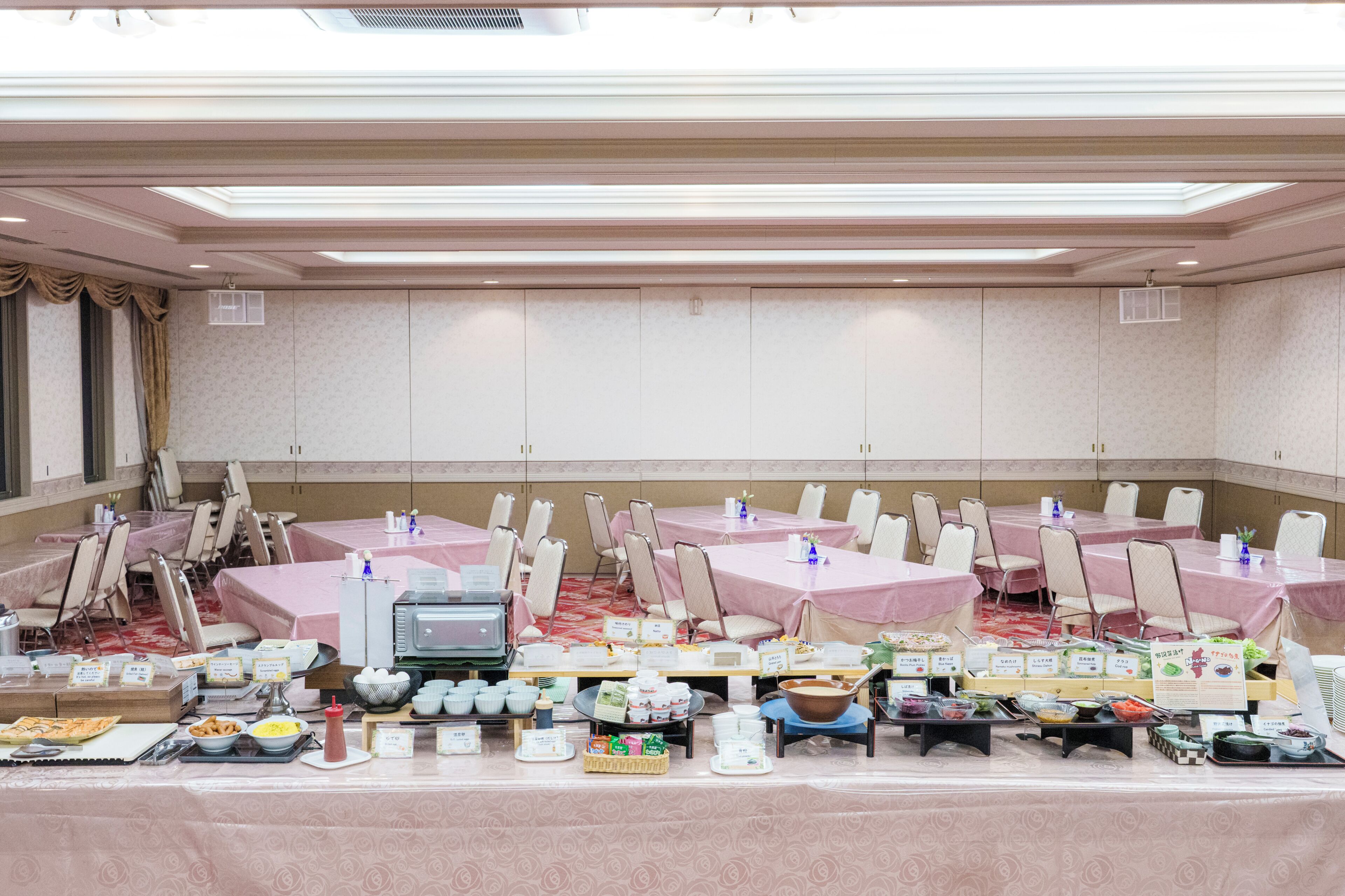 A banquet hall with tables covered in pink tablecloths, chairs, and a buffet setup with various food items.