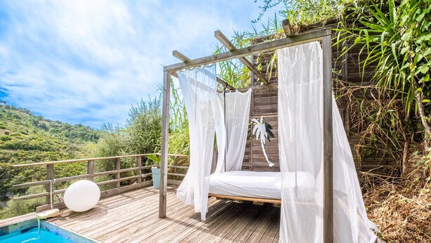 Eine Holzterrasse mit einem Bett unter einem Baldachin, einem Pool und Blick auf einen grünen Hügel.