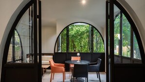 A room with a round table and chairs, a bench, and large arched windows with a view of greenery. - O2 The Residence (Kanchanaburi)
