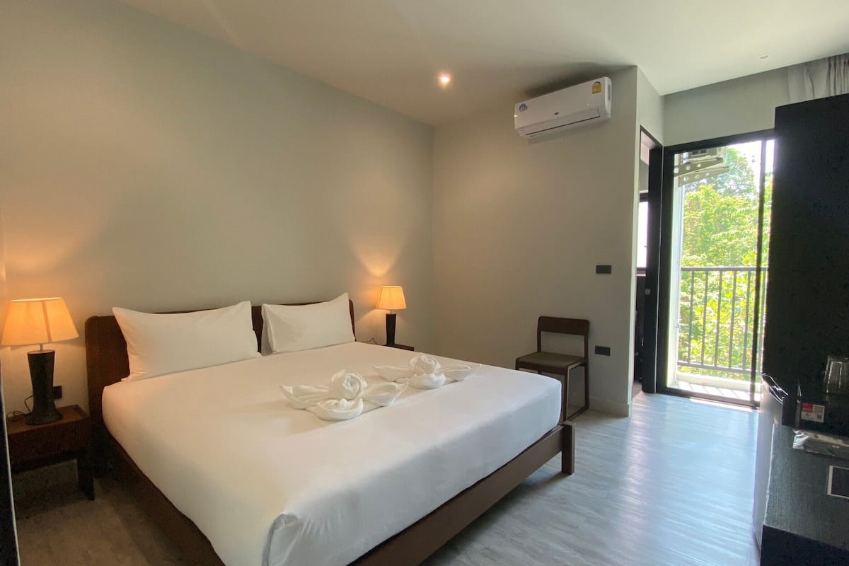 A hotel room with a large bed, two bedside lamps, an air conditioning unit, a chair, and a balcony with a view of greenery.