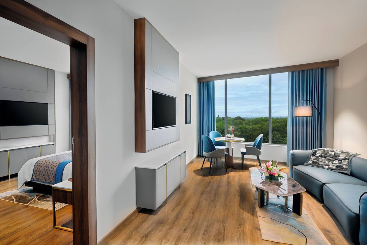 A modern hotel room with a large bed, a TV mounted on the wall, a dining area with a table and chairs, and a sofa.