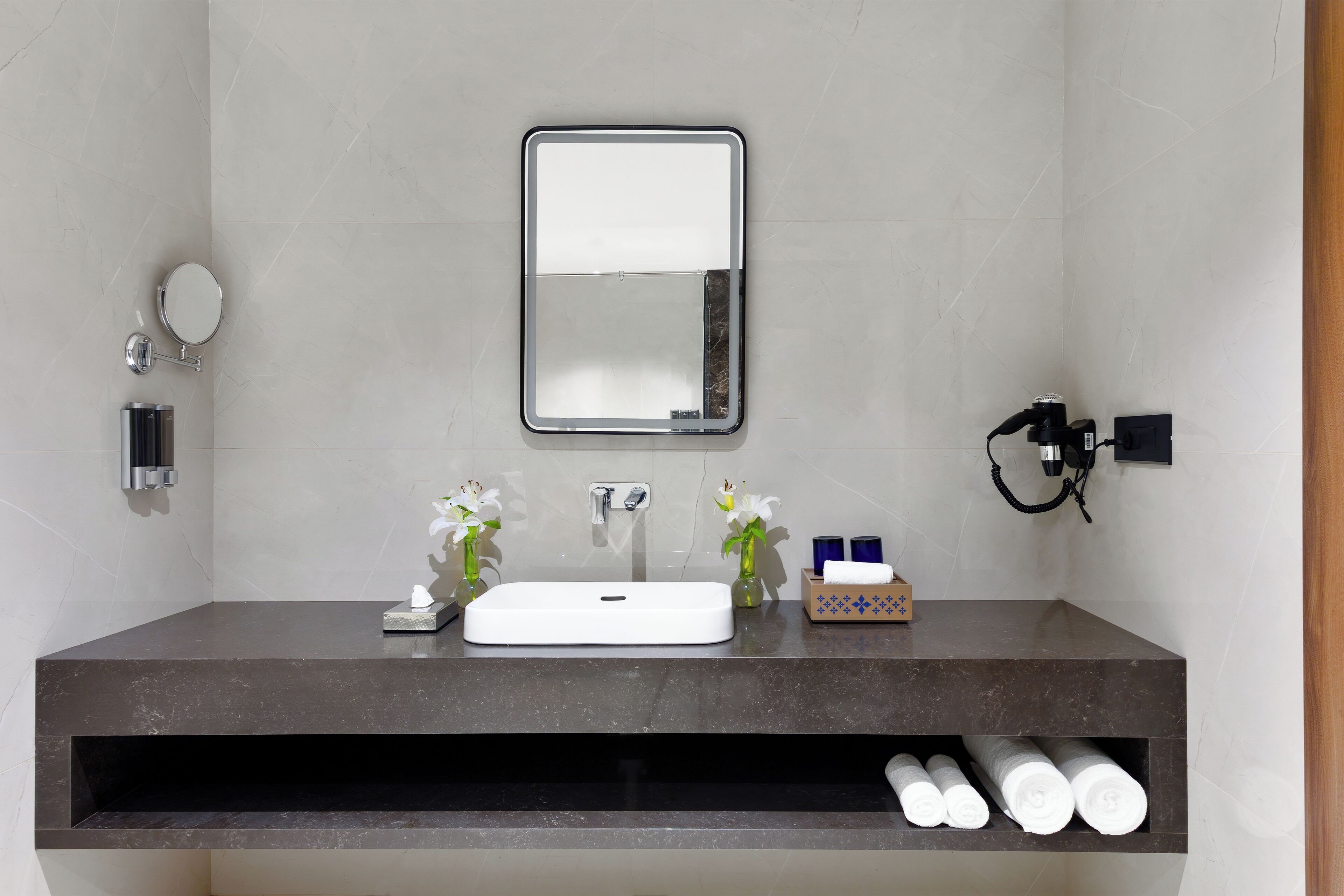 A modern bathroom with a dark countertop, a white sink, a mirror, and two vases with white flowers.