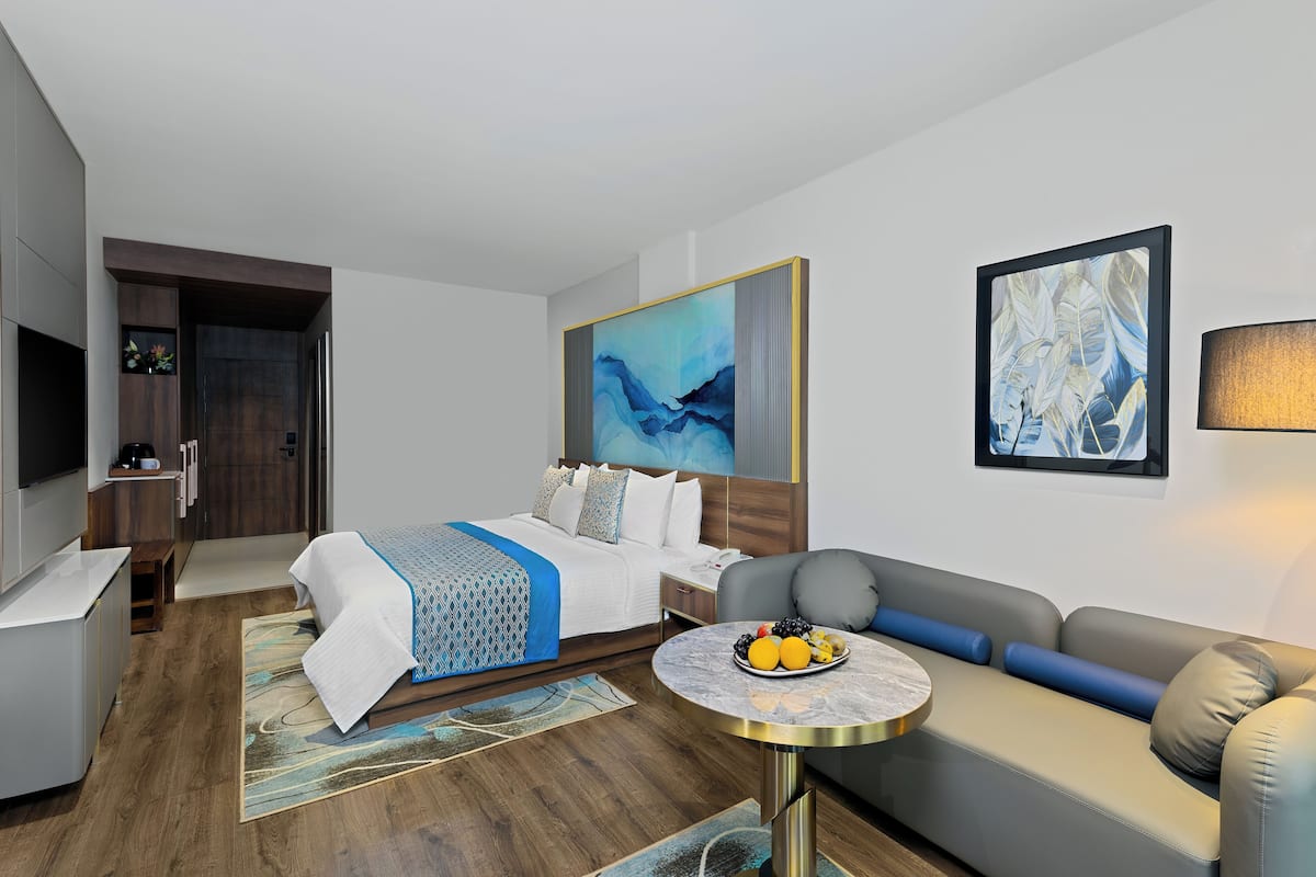A modern hotel room with a bed, a sofa, a small table with fruit, a TV, and artwork on the walls.
