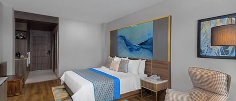 A modern hotel room with a large bed, a sitting area with a chair, a desk, and a small kitchen area.