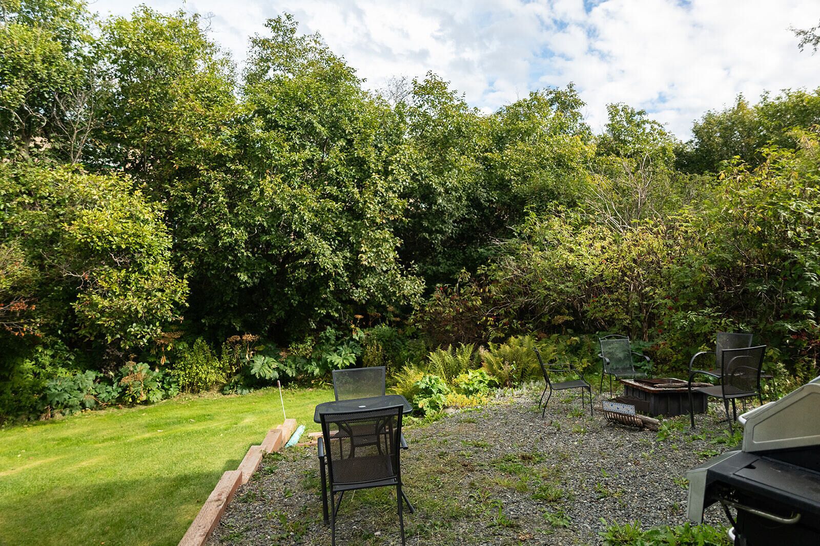 A backyard with a lawn, a grill, and metal chairs.