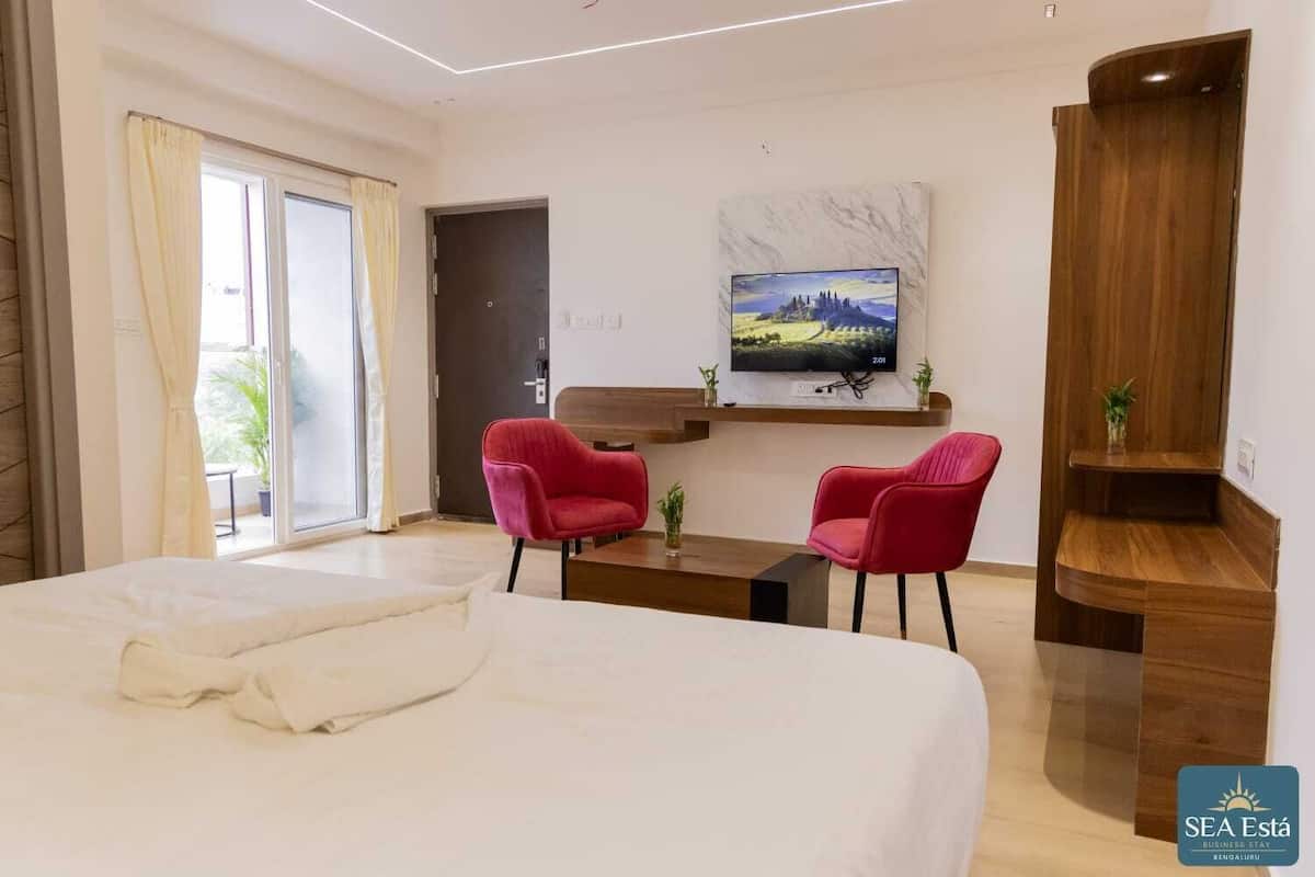 Una habitación de hotel moderna con una cama grande, dos sillas rojas, un televisor en la pared y un aparador de madera.