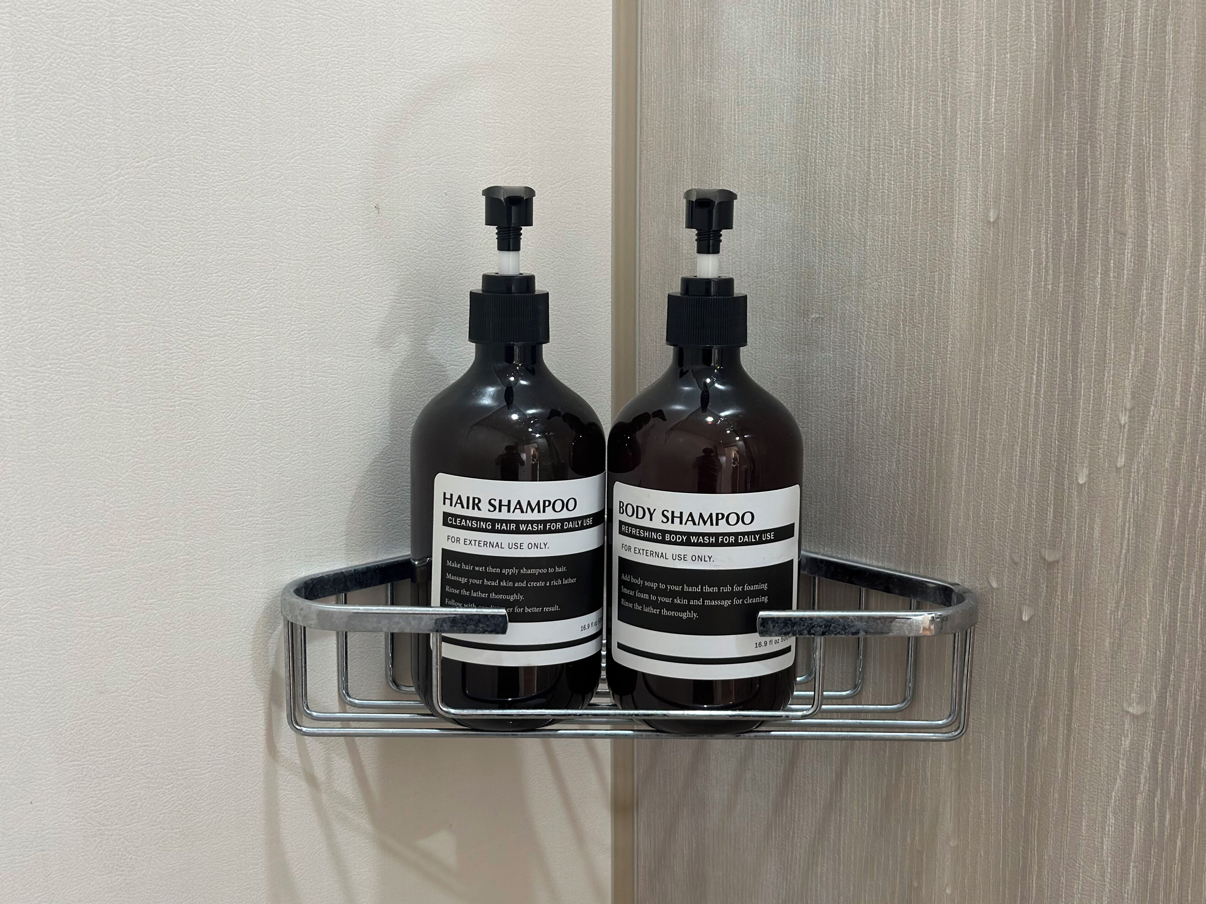 Twee flessen shampoo voor haar en lichaam op een metalen plank tegen een muur.
