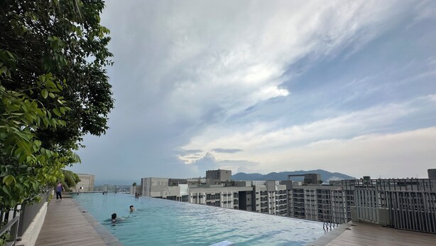 Una piscina infinita en la azotea con terraza de madera, rodeada de edificios y árboles, con vistas a montañas lejanas.