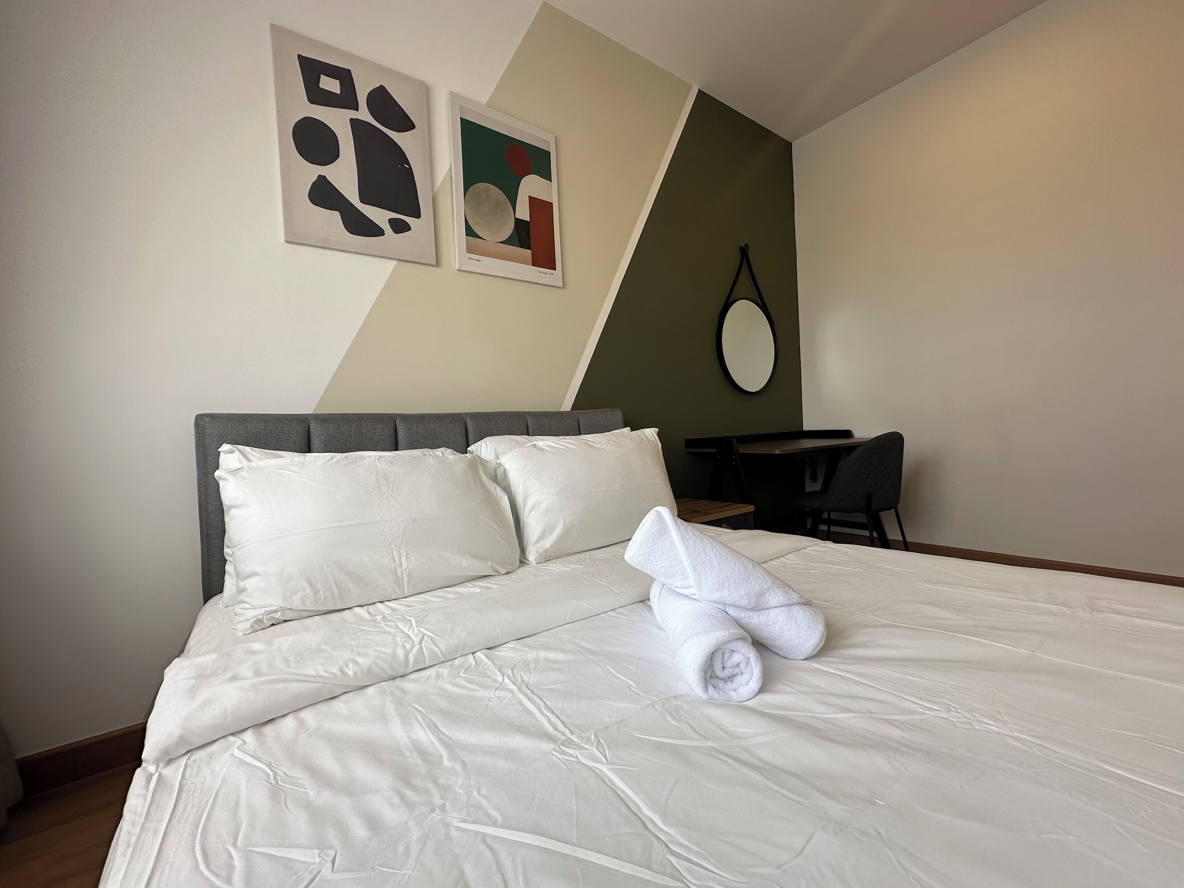 A neatly made bed with white linens and a rolled towel, a small desk with a chair, and two abstract paintings on the wall.