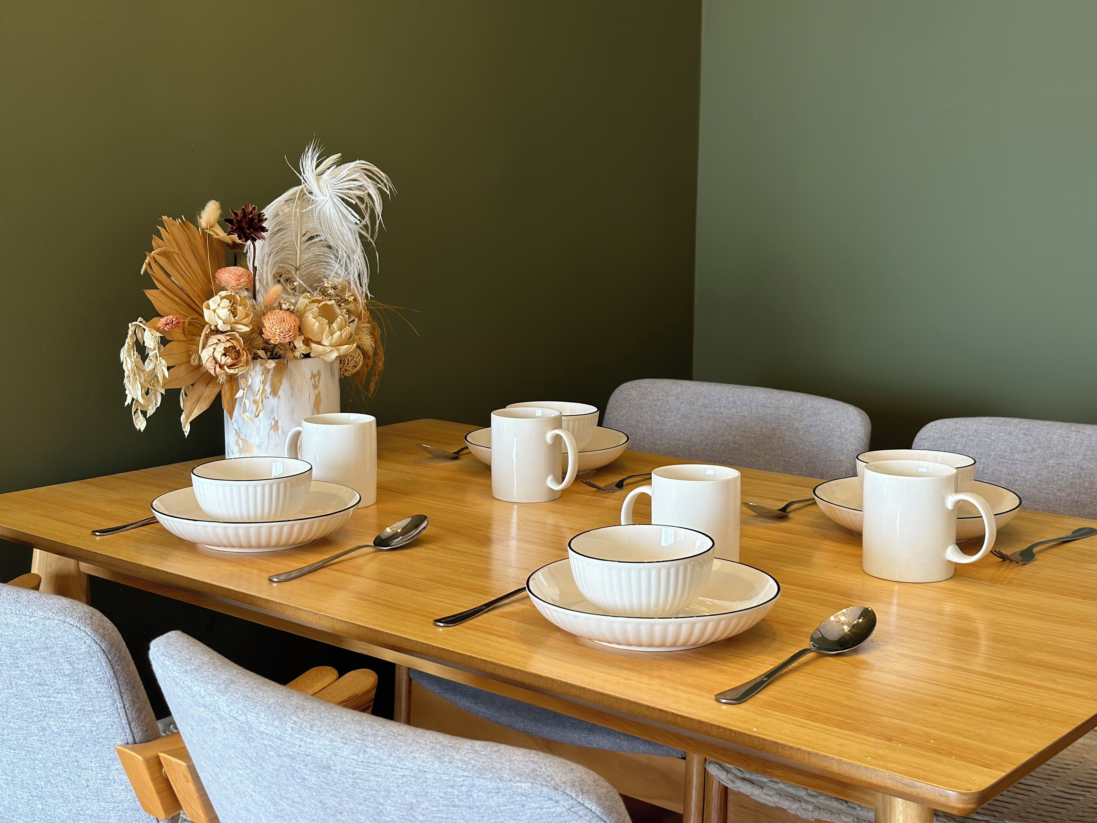 A dining table set with white ceramic dishes, bowls, and mugs, accompanied by a vase of dried flowers.
