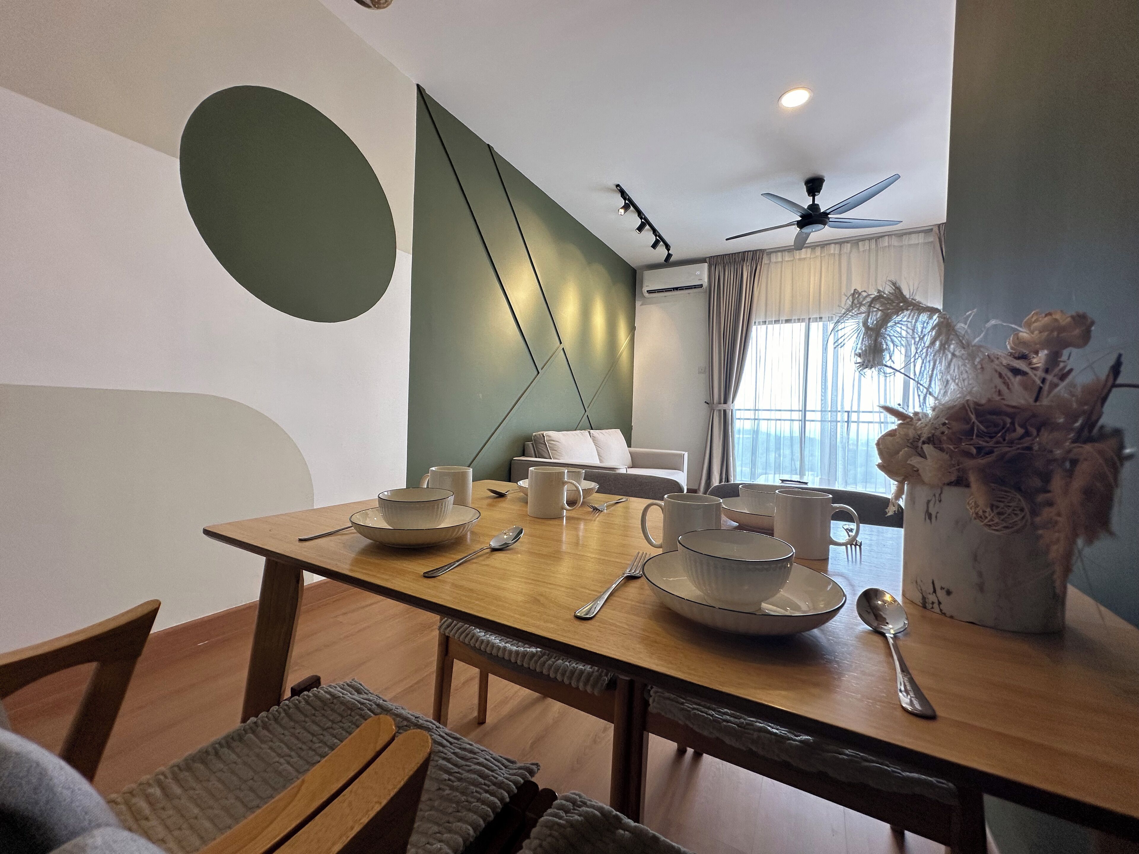A dining area with a wooden table set for four, a sofa in the background, and a decorative vase with dried flowers.