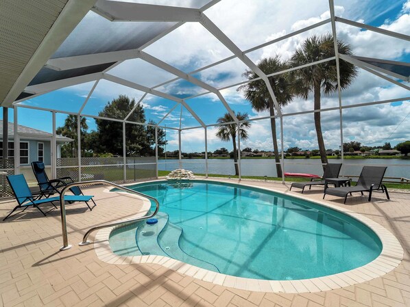 A covered outdoor swimming pool with lounge chairs, a view of a lake, and palm trees.