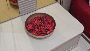 Une table de chevet blanche avec un bouton doré, un miroir reflétant un canapé rouge et un coussin à motifs floraux, et un bol rempli de fleurs rouges séchées posé sur la table.