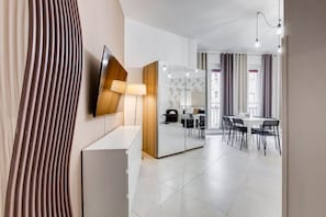 A modern hotel room with a flat-screen TV, a mirror, a desk with a lamp, a dining area with chairs, and a view of the street.
