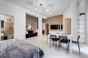 A modern hotel room with a bed, a dining area with chairs and a table, a sofa, a TV, and a bathroom with a mirror and sink.