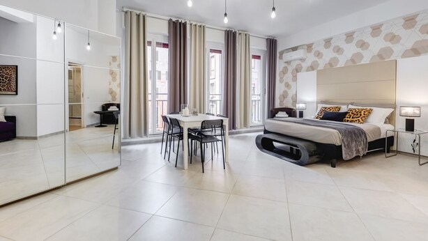 Una habitación de hotel moderna con una cama grande, una zona de comedor con una mesa y sillas, un espejo amplio y vistas a la ciudad desde las ventanas.