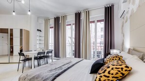 Una habitación de hotel moderna con cama, escritorio y sillas, un balcón con cortinas y vistas a la ciudad.