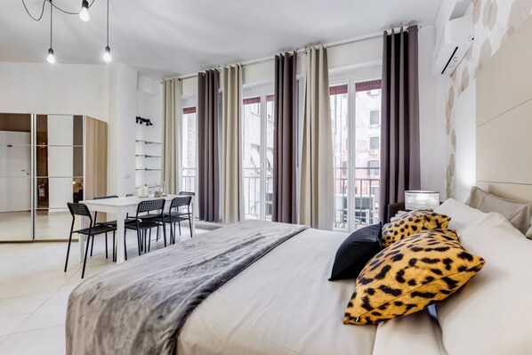 A modern hotel room with a bed, a desk, and chairs, a balcony with curtains, and a city view.