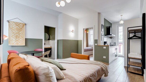 Una habitación moderna y luminosa con cama, una pequeña mesa de comedor, una zona de cocina y un vestidor.