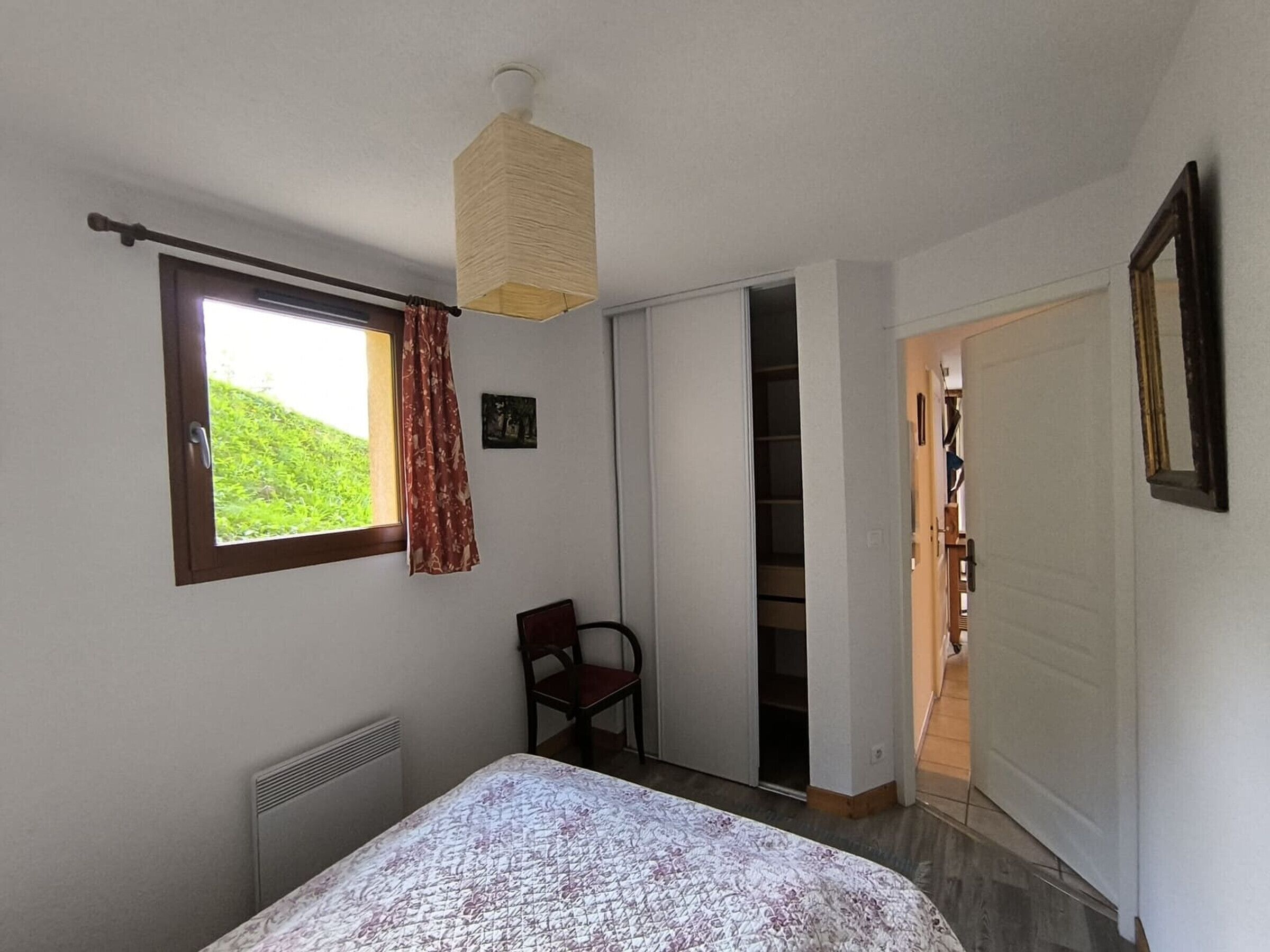 Een slaapkamer met een bed, een raam met gordijnen, een stoel, een kast en een deur naar een andere kamer.