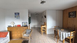 Een gezellige woonkamer met een houten dressoir, een rieten fauteuil, een eettafel met stoelen, en een keuken op de achtergrond.