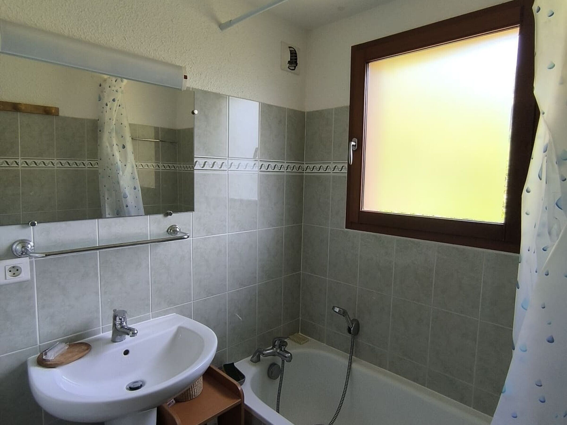 A bathroom with a sink, mirror, shower, and a window with a curtain.
