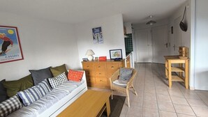 Een woonkamer met een bank, een houten tafel, een rieten stoel, een dressoir en een deur die naar de keuken leidt.