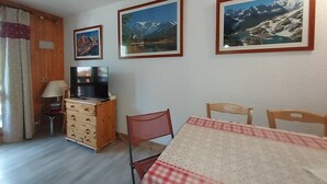 A room with a bed, a TV, a wooden dresser, a table with a checkered tablecloth, and three framed pictures on the wall.