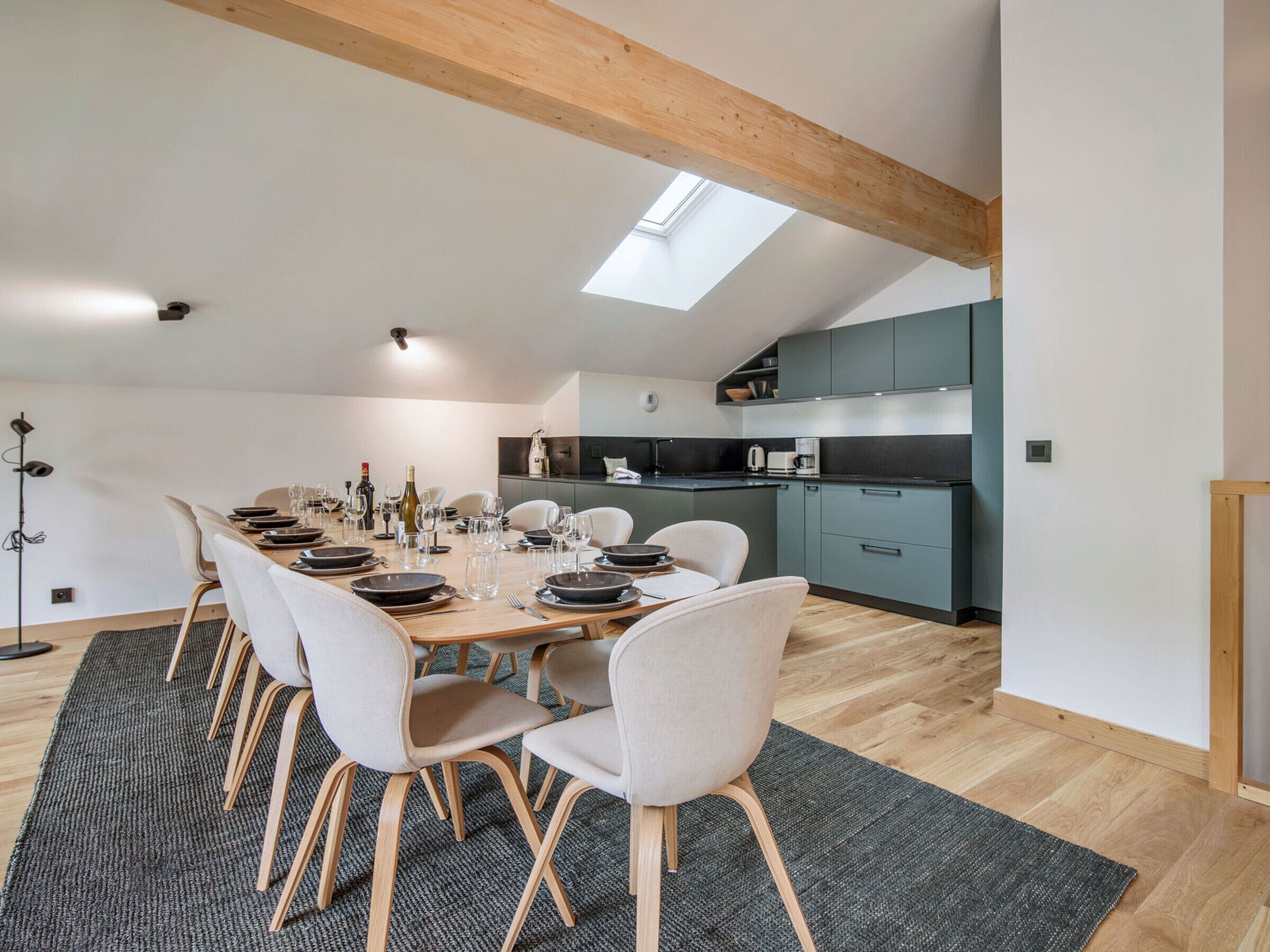 Un espace repas moderne avec une longue table dressée pour six personnes, des chaises de couleur claire et une lucarne au-dessus.