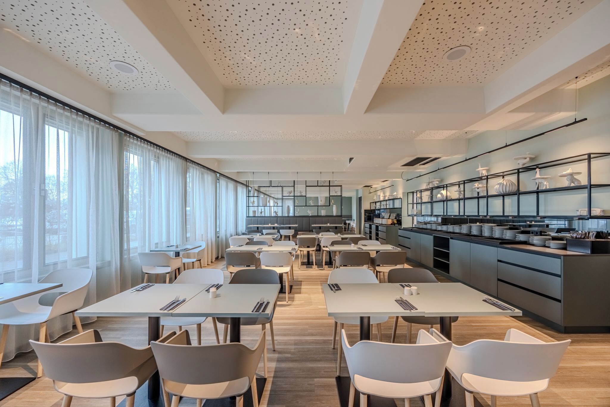 A modern dining area with tables and chairs, a kitchen area with shelves, and large windows with curtains.