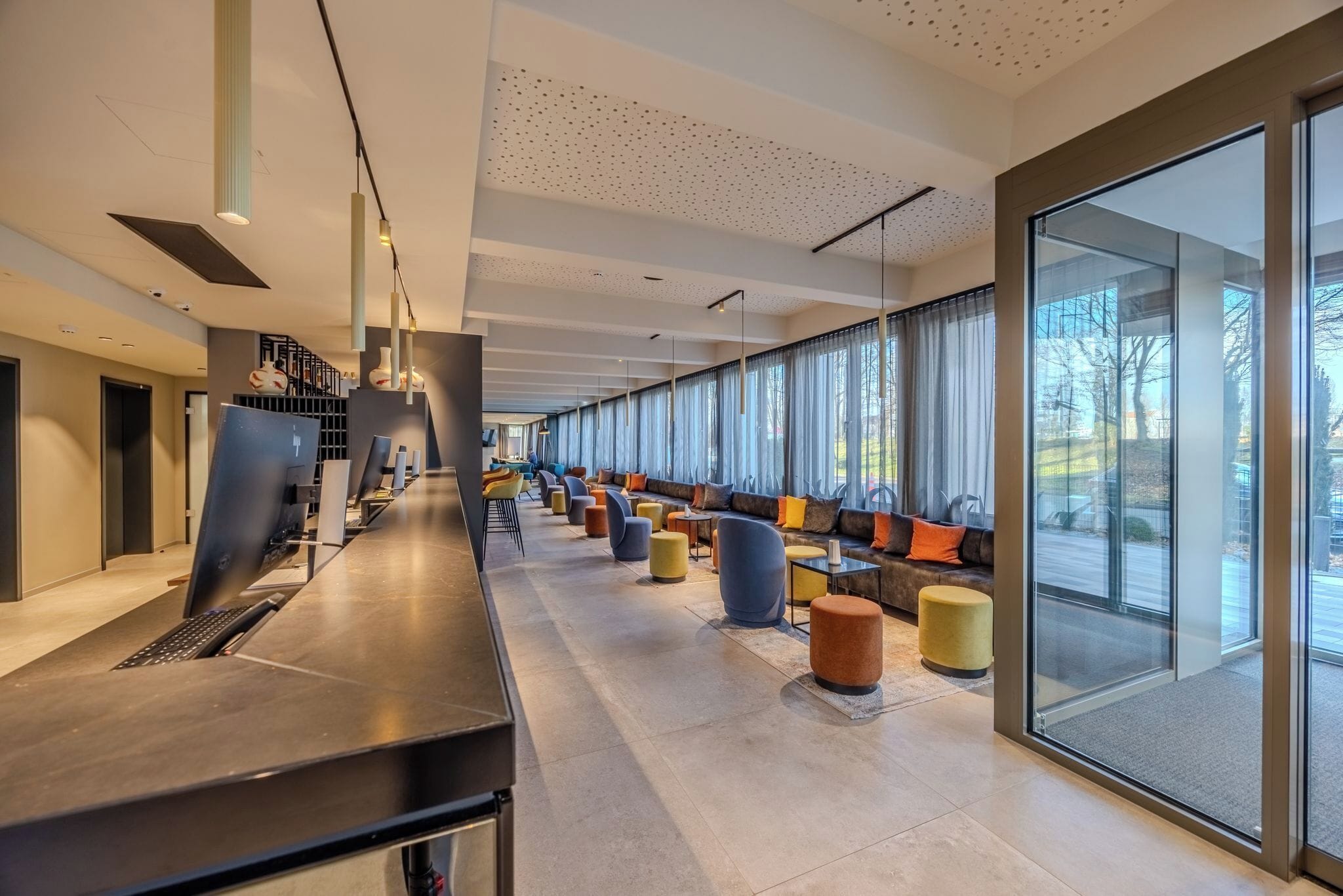 A modern hotel lobby with a long reception desk, seating areas with colorful ottomans, and large windows offering views of the outdoors.