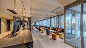 A modern hotel lobby with a long reception desk, seating areas with colorful ottomans, and large windows offering views of the outdoors.