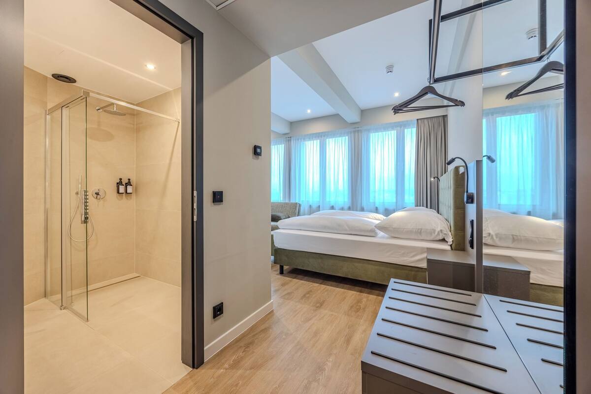 Une chambre d’hôtel moderne dotée d’une douche en verre, d’un lit avec du linge de maison blanc et d’un grand miroir avec un porte-manteau.