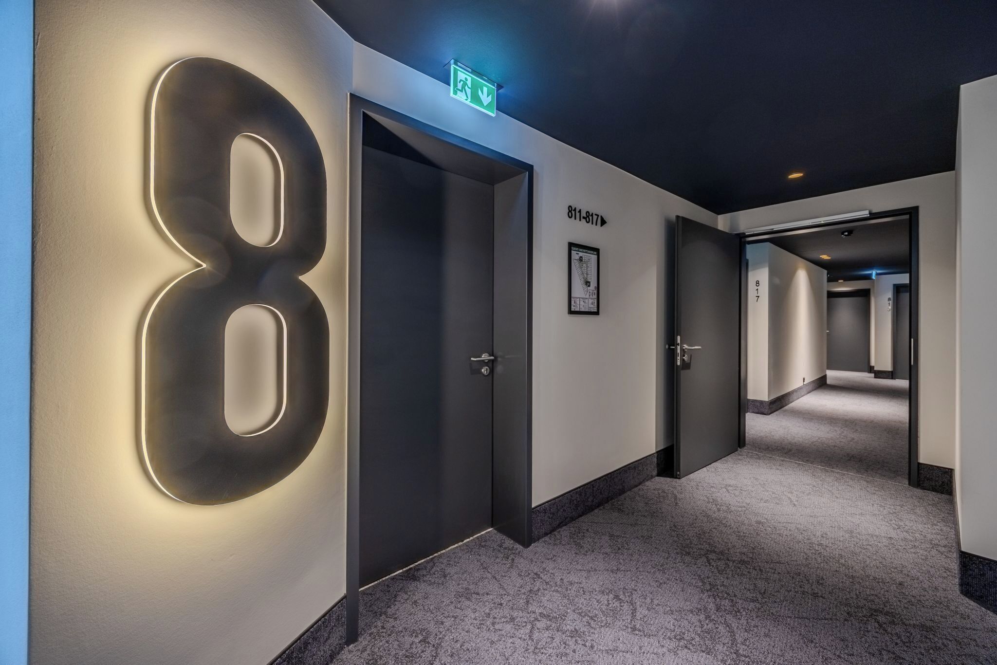 A hallway with a large illuminated number '8' on the wall, an emergency exit sign above a door, and a framed picture on the wall.