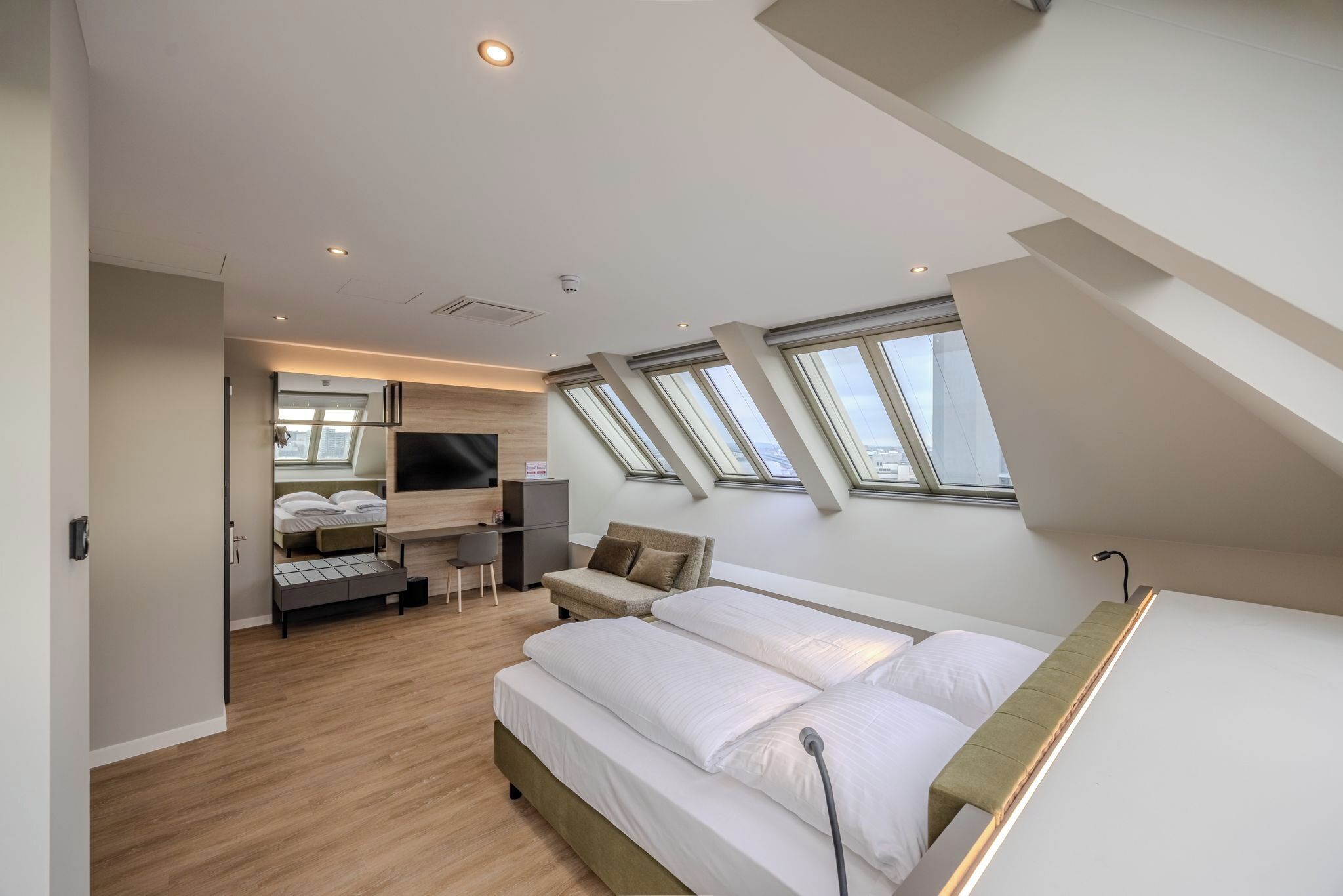 A modern hotel room with a lofted ceiling, skylights, a bed, a seating area, and a TV.