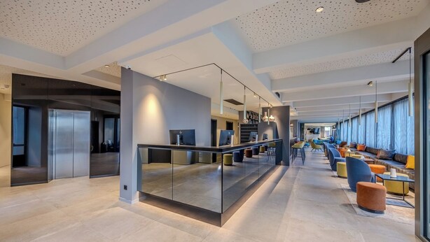 Un hall d’entrée spacieux au design moderne, doté d’une cloison en verre, d’un espace salon avec des fauteuils colorés et d’un long comptoir équipé d’ordinateurs.