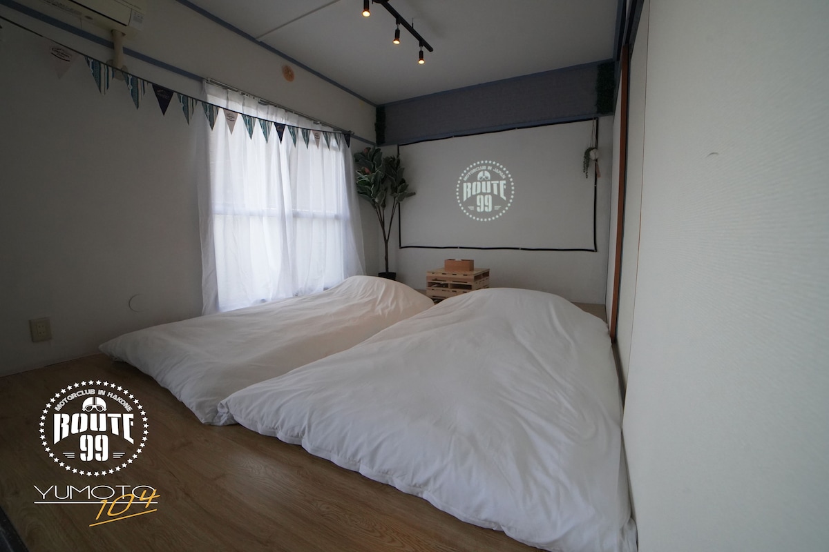 A room with two beds, a window with sheer curtains, a wall-mounted projector screen, and a wooden floor.