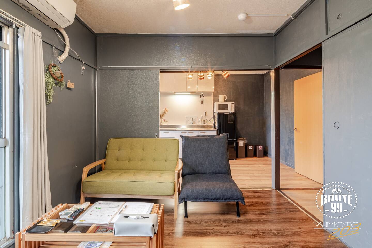 Un espacio habitable compacto con un sofá, una silla, una mesita con objetos y una kitchenette al fondo.