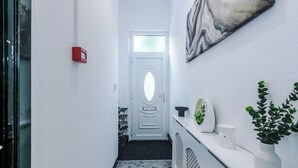 A narrow hallway with a white door, a small red fire alarm, a marble-patterned wall, and a potted plant on a white shelf.