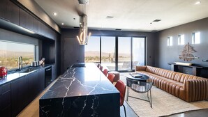 A modern kitchen with a marble island, a dining area with a leather sofa, and large windows offering a view of the desert. - Black Casita by Fieldtrip - Bold Villa w Pool + Spa (Joshua Tree)