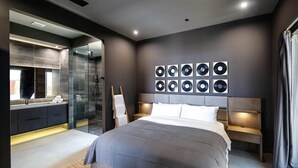A modern bedroom with a large bed, a glass shower enclosure, a wall of vinyl records, and a dark color scheme. - Black Casita by Fieldtrip - Bold Villa w Pool + Spa (Joshua Tree)