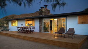 A modern house with a stone chimney, a patio with a table and chairs, and a large sliding glass door leading to an interior space.