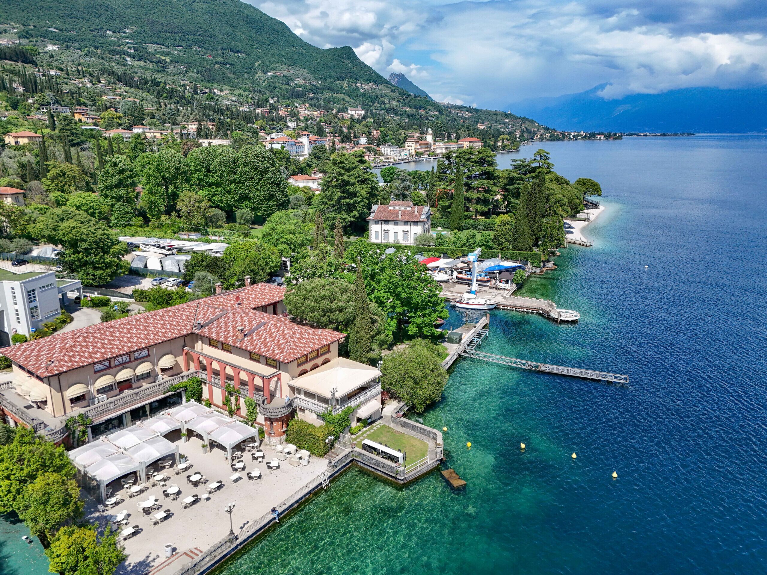 An aerial view of a coastal town with a prominent building featuring a red-tiled roof, a waterfront promenade, and clear blue waters.