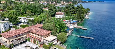 An aerial view of a coastal town with a prominent building featuring a red-tiled roof, a waterfront promenade, and clear blue waters.