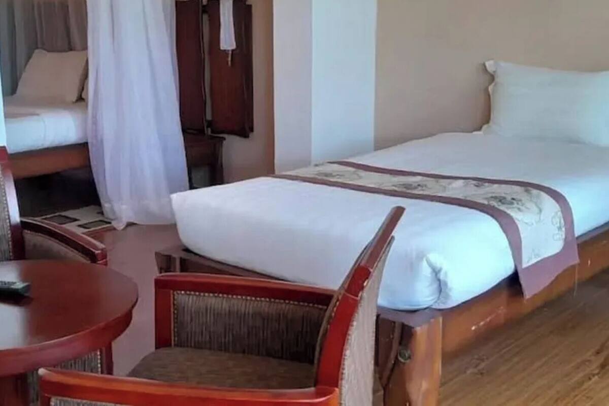 Una cama individual con colchón blanco y cabecera marrón, una mesita de noche de madera con lámpara, una silla roja y una pequeña mesa redonda con un control remoto.