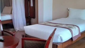 A single bed with a white mattress and a brown headboard, a wooden nightstand with a lamp, a red chair, and a small round table with a remote control.
